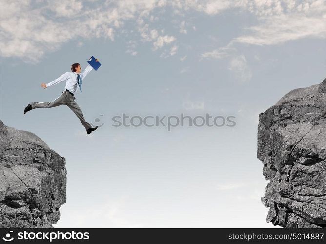 Brave businessman. Young businessman with suitcase jumping over mountain gap