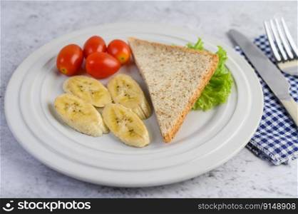 bread, banana, and tomato on white plate with fork and a knife.