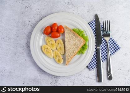 bread, banana, and tomato on white plate with fork and a knife.