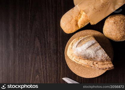 bread concept a half loaf of bread on the wooden plate, a loaf of bread into a paper bag, a knife, and a bun put on a black wooden scene.