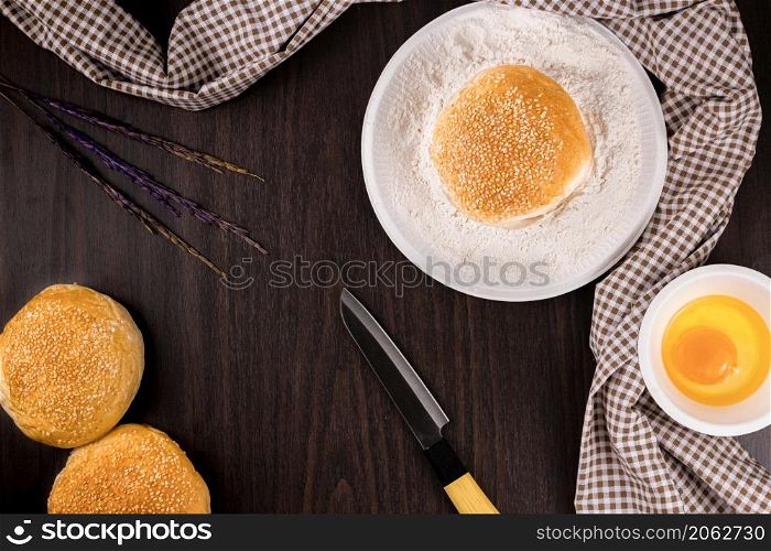 bread concept buns, a dish of flour, a knife, and a tiny bowl of raw egg lying on the dark wooden table.
