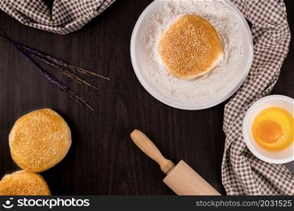 bread concept buns, a dish of flour, a rolling pin, and a tiny bowl of raw egg lying on the dark wooden table.