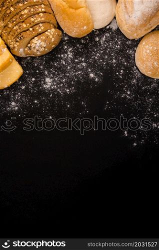 bread concept several slices of dark brown bread with seeds, buns, croissants, and loaves of bread gathered together on the black background with sprinkling flour dust.