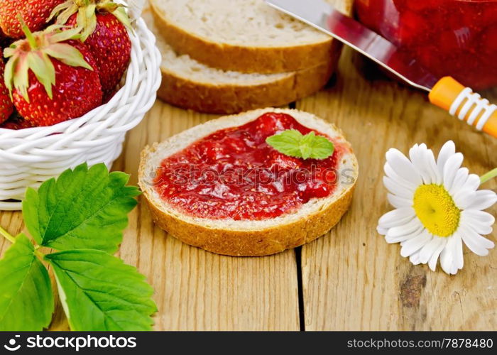 Bread with strawberry jam, a jar of jam, knife, strawberries in a wicker basket, chamomile on a wooden boards background