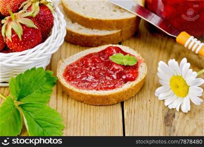 Bread with strawberry jam, a jar of jam, knife, strawberries in a wicker basket, chamomile on a wooden boards background