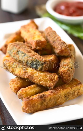 Breaded fried zucchini sticks with ketchup in the back, photographed with natural light (Selective Focus, Focus on the front of the zucchini stick in the middle of the image)