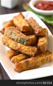 Breaded fried zucchini sticks with ketchup in the back, photographed with natural light (Selective Focus, Focus on the front of the zucchini stick in the middle of the image)