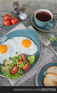 Breakfast consists of fried eggs, bacon and various vegetables (tomato, green pea, broccoli, lettuce), toast and a cup of coffee on the old wooden table; vertical image