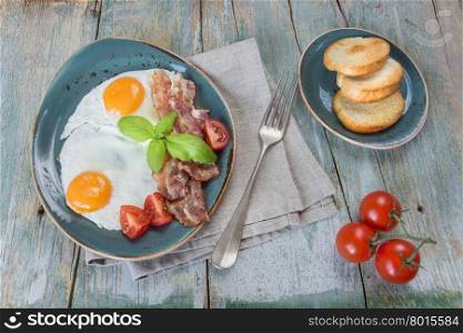 Breakfast consists of fried eggs, bacon, tomato and toast on the old wooden table
