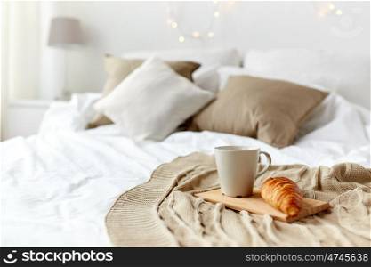 breakfast, coziness, morning, holidays and winter concept - cozy bedroom with coffee cup and croissant on wooden board in bed at home