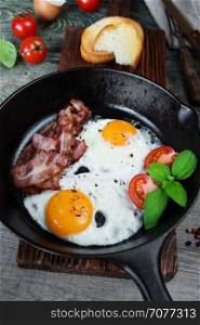 Breakfast in a rustic style: two fried eggs, strips of bacon, fresh tomatoes and basil in a cast iron skillet and toasts on an old wooden table