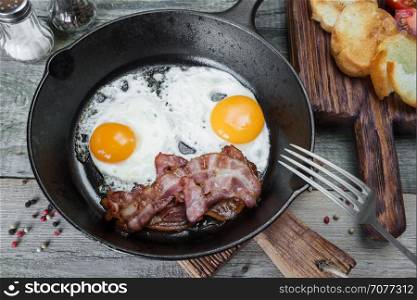 Breakfast in a rustic style: two fried eggs, strips of bacon in a cast iron skillet and toasts on an old wooden table