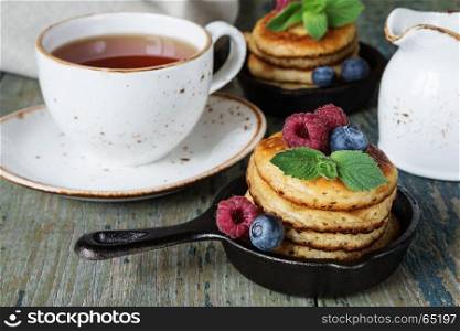 Breakfast of pancakes in a portioned cast-iron frying pan, fresh berries and black tea in a vintage ceramic cup on an old wooden table