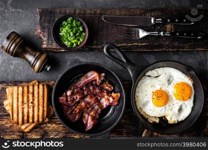 breakfast or brunch fried bacon and eggs in black skillets and crispy toasts top view