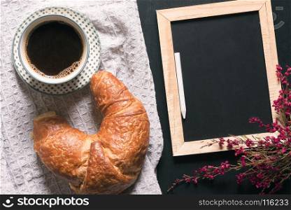 Breakfast table with a cup of coffee and a croissant on a vintage kitchen towel, near an empty chalkboard surrounded by flowers, on a black table.