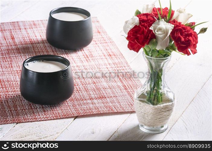 Breakfast table with two black mugs full of fresh milk and a bouquet of white roses and red carnations, on a white table, on a sunny morning.