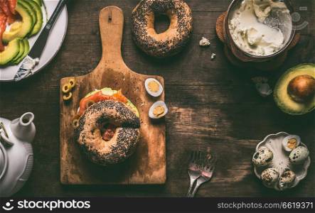 Breakfast with bagel bread topped with salmon, avocado and fresh cheese on dark rustic wooden background, top view. Copy space for your design