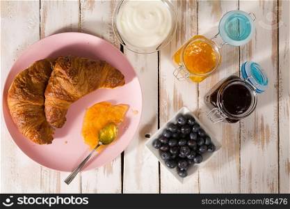 Breakfast with croissant, blueberries, yogurt, orange and blueberry jam over a wooden table seen from above. Breakfast with croissant and blueberries over a wooden table