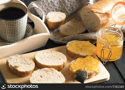 Breakfast with french bread, sliced on a wooden cutting board, and smeared with homemade peach jam and a cup of hot coffee on a white wooden tray.