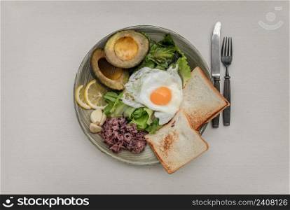 Breakfast with Fried Egg to put it on top of Fresh vegetables, Rice berry, Breads, and Garlic with Avocado cut in half and lemon sliced on Ceramic plate. Healthy food concept, Top view, Selective Focus.