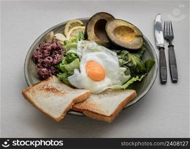 Breakfast with Fried Egg to put it on top of Fresh vegetables, Rice berry, Breads, and Garlic with Avocado cut in half and lemon sliced on Ceramic plate. Healthy food concept, Selective Focus.