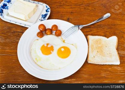 breakfast with two fried eggs on white plate, cherry tomatoes, fresh toast and dairy butter in butterdish on wooden table
