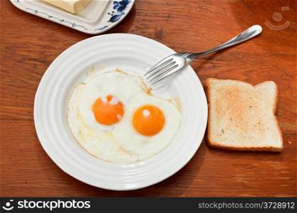 breakfast with two fried eggs on white plate, fresh toast and dairy butter in butterdish on wooden table
