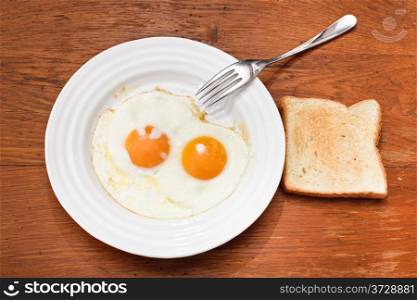 breakfast with two fried eggs on white plate, fresh toast on wooden table