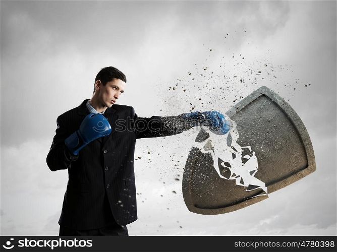 Breaking all doubts. Young businessman in blue boxing gloves breaking stone shield