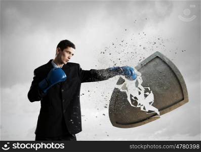 Breaking all doubts. Young businessman in blue boxing gloves breaking stone shield