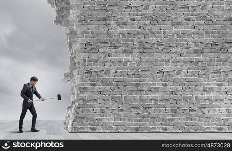 Breaking barriers. Young determined businessman breaking wall with hammer