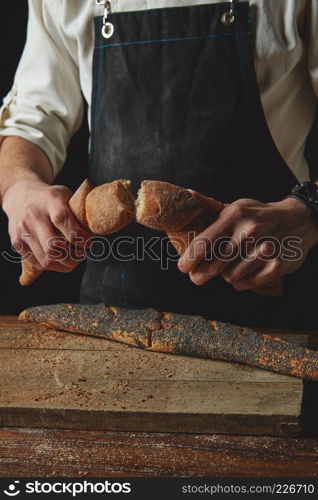 Breaking fresh bread. Baking and cooking concept background. Hands tearing apart loaf on rustic wooden table. Hand breaks baguette