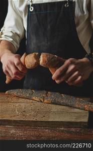Breaking fresh bread. Baking and cooking concept background. Hands tearing apart loaf on rustic wooden table. Hand breaks baguette