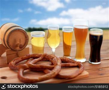 brewery, alcohol drinks and food concept - different types of beer in glasses, wooden barrel and pretzel on table over cereal field and blue sky background. beer glasses, barrel and pretzel over cereal field