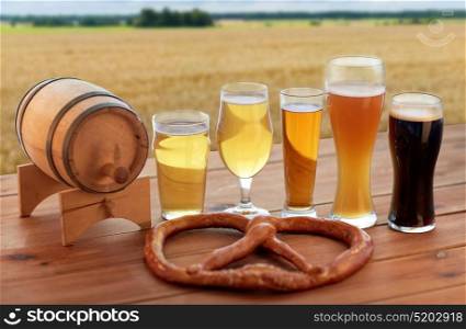 brewery, alcohol drinks and food concept - different types of beer in glasses, wooden barrel and pretzel on table over cereal field background. beer glasses, barrel and pretzel over cereal field