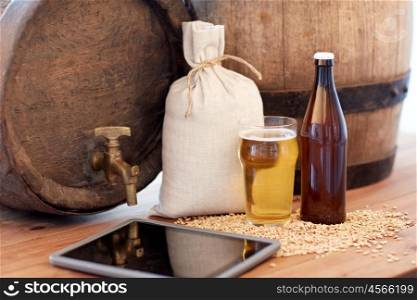 brewery, drinks and alcohol concept - close up of old beer barrel, glass bottle, tablet pc computer and bag with malt on wooden table