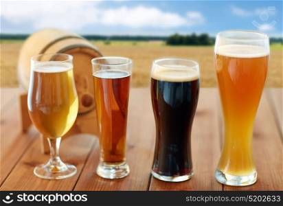 brewery, drinks and alcohol concept - different types of beer in glasses and barrel on table over cereal field and blue sky background. different types of beer in glasses on table