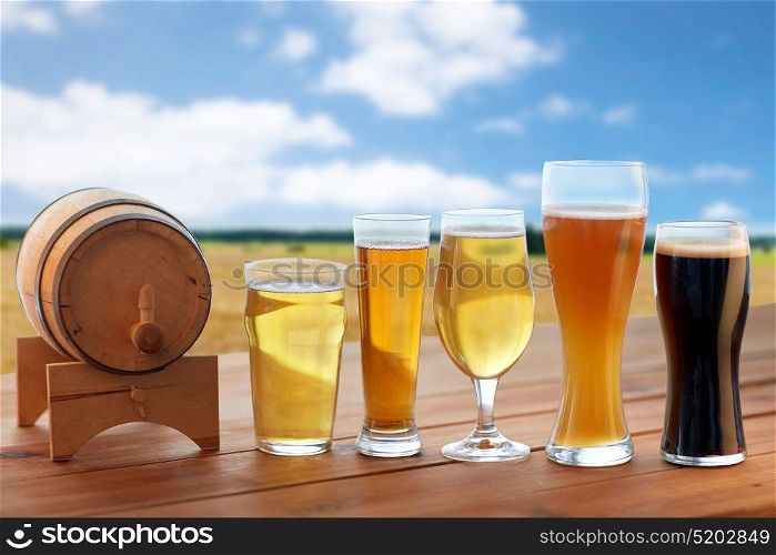 brewery, drinks and alcohol concept - different types of beer in glasses and barrel on table over cereal field and blue sky background. different types of beer in glasses on table