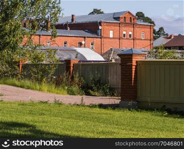 Brick house behind a fence with outbuildings on a sunny day.