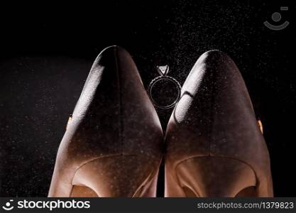 Bridal wedding shoes and ring with diamond. Wedding ring between beige bridal shoes on dark background. Wedding women accessories concept. Bridal wedding shoes and ring with diamond. Wedding ring between beige bridal shoes on dark background. Wedding women accessories concept.