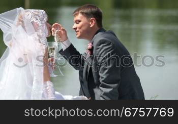 bride and groom drink champagne sitting on the shore of Lake