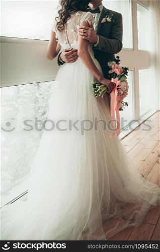 Bride and Groom with a wedding bouquet standing by the big window, holding each other in arms, view from the back