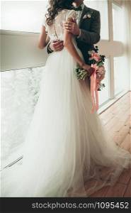 Bride and Groom with a wedding bouquet standing by the big window, holding each other in arms, view from the back