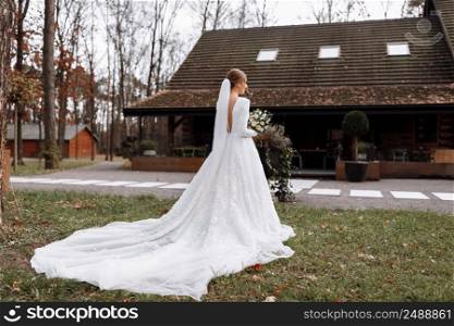 bride in a long white dress is standing in the park and holding a wedding bouquet of roses flowers and greens. young woman with hairstyle, makeup and long veil outdoors near hotel.. bride in a long white dress is standing in the park and holding a wedding bouquet of roses flowers and greens. young woman with hairstyle, makeup and long veil outdoors near hotel