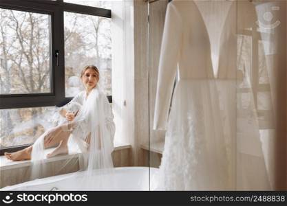 Bride s morning. Bride drinking champagne in the peignoir. young woman is sitting on a large window in a hotel room in bathroom. Beautiful girl in white wedding robe. wedding day.. Bride s morning. Bride drinking champagne in the peignoir. young woman is sitting on a large window in a hotel room in bathroom. Beautiful girl in white wedding robe. wedding day