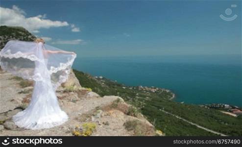 bride stands on cliff with a flowing veil on the wind against the blue sea and sky
