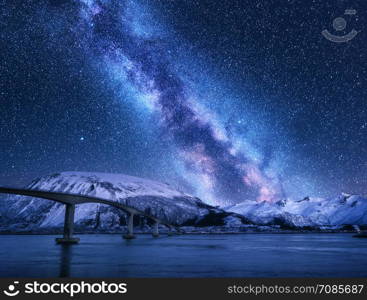 Bridge and starry sky with Milky Way over snow covered mountains reflected in water. Night landscape with road, snowy rocks, purple sky with stars and milky way, sea. Winter in Lofoten islands, Norway