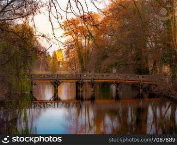 Bridge over a dark small lake with a red mill shining in the evening sun in the background