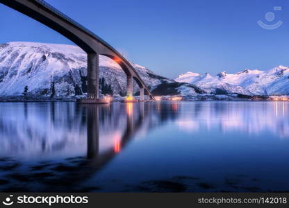 Bridge with illumination, snow covered mountains, village and blue sky with beautiful reflection in water. Night landscape with bridge, snowy rocks reflected in sea. Winter in Lofoten islands, Norway