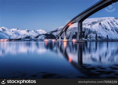 Bridge with illumination, snow covered mountains, village and blue sky with reflection in water. Night landscape with bridge, snowy rocks reflected in sea. Winter in Lofoten islands, Norway. Roadway